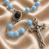 A close-up of a rosary with light blue beads, a silver-tone medallion engraved 'TERRA JERUSALEM', and a silver crucifix, on a beige satin fabric background.