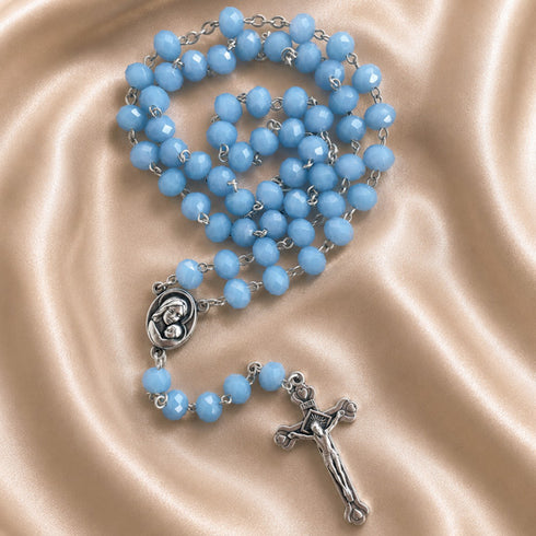 A light blue rosary with silver-tone metal links, a round medallion, and a crucifix, on a draped satin fabric background.