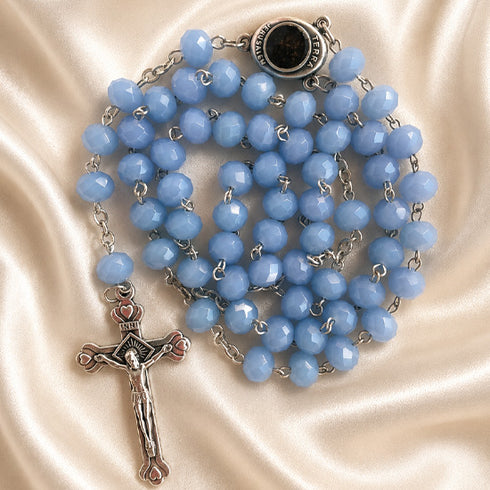 A light blue faceted bead rosary with silver-tone metal links, a round dark centerpiece, and a silver crucifix, on a satin background.