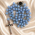 A light blue faceted bead rosary with silver-tone metal links, a round dark centerpiece, and a silver crucifix, on a satin background.