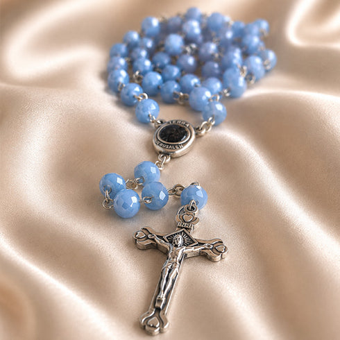 A rosary necklace with light blue faceted beads and a silver-tone crucifix pendant, displayed on draped champagne satin fabric.