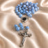 A rosary necklace with light blue faceted beads and a silver-tone crucifix pendant, displayed on draped champagne satin fabric.