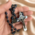 A close-up of a hand holding a dark blue and silver beaded rosary with a silver crucifix, on a beige satin fabric.