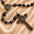 A close-up of a rosary on a satin fabric background. The rosary with dark beads, a metallic silver spacer beads and antique bronze-toned.