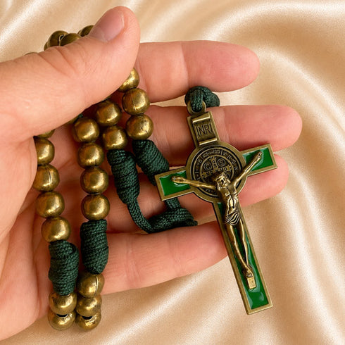 A close-up of a hand holding a St. Benedict rosary with green and gold beads and a crucifix, on a satin fabric.