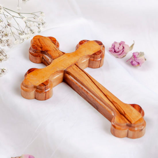 Small handmade olive wood Eastern cross with carved edges, photographed on white with rose petals.