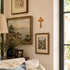Decorative wall with framed landscape paintings and a cross, near a window.
