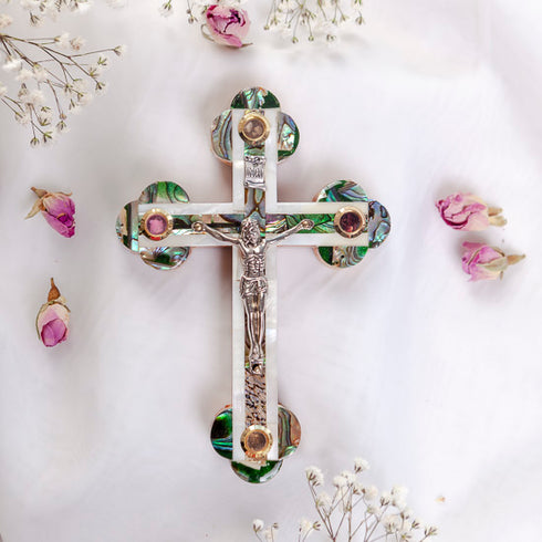 Budded olive wood cross with crucifix, white and green Mother of Pearl inlay, lying flat on white background with petals.