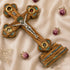 Wooden cross with intricate designs and a figure on a beige fabric background with dried flowers.