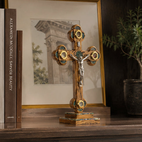 Decorative cross with gold and green accents on a wooden surface, with books and a framed picture in the background.