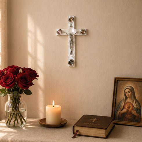 Wall cross, candle, vase of red roses, and a Bible on a table with a religious painting in the background.
