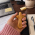 A hand with pink nails holds a small, polished palm cross. The background shows a desk with a laptop, notebook, and pen.
