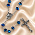 Blue crystal rosary with silver-tone Virgin Mary centerpiece and ornate crucifix displayed on champagne satin.