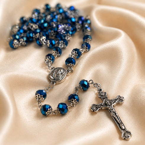 A close-up of a rosary with faceted blue beads, centerpiece medallion, and crucifix, arranged on champagne satin fabric.
