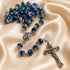 A close-up of a rosary with faceted blue beads, centerpiece medallion, and crucifix, arranged on champagne satin fabric.