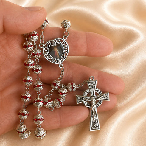 A close-up of a hand holding a silver rosary with red enamel accents and a small religious medallion.