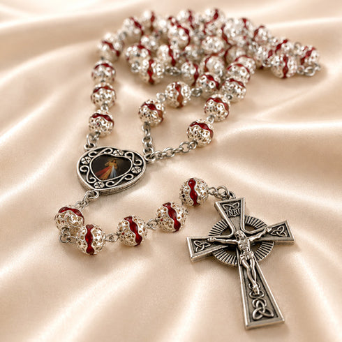 Silver rosary with red and clear beads, a silver crucifix, and a central medallion, on a beige satin fabric background.