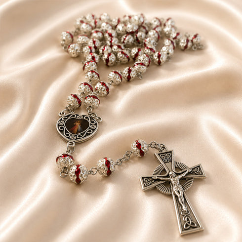 A silver-toned rosary with red enamel accents and a crucifix pendant, displayed on draped cream satin fabric.