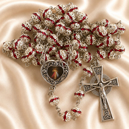A silver-toned rosary with red enamel accents, featuring a central heart-shaped medallion with a religious image and a crucifix.