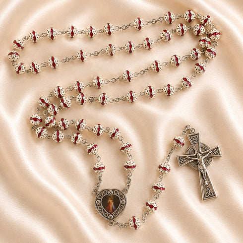 Silver rosary with red and white beads, a heart-shaped pendant and a silver crucifix, on a beige satin fabric background.