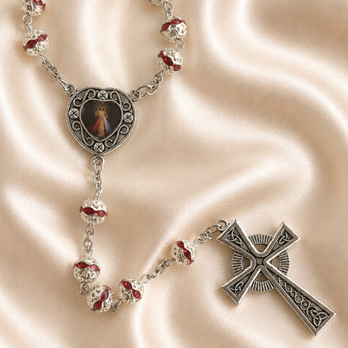 Silver rosary with red enamel beads and a central medallion featuring a religious image, on a beige fabric background.