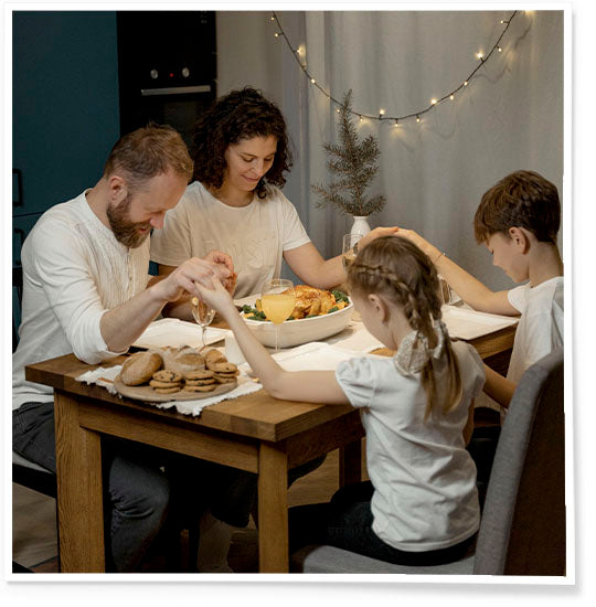 Family dressed in white holding hands and praying over a table with food on it.