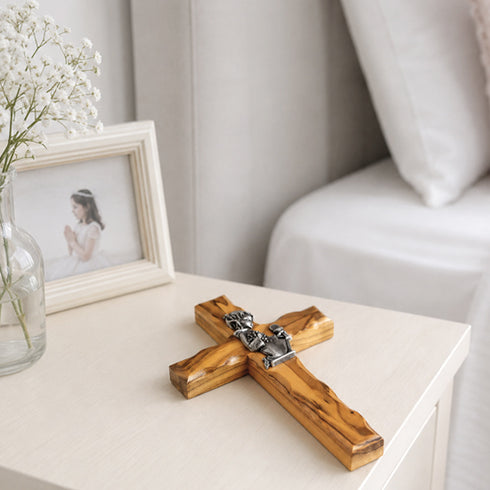 Wooden cross with silver details on a white surface, next to a photo frame and vase with flowers.