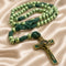 A green beaded rosary with knotted cords and a gold-toned crucifix, on a beige satin fabric.
