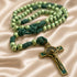 A green beaded rosary with knotted cords and a gold-toned crucifix, on a beige satin fabric.