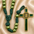 A green and gold rosary with green beads and a green cross pendant, displayed on a beige satin fabric background.