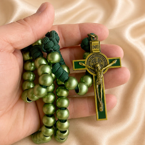 A hand holds a green and gold St. Benedict Rosary against a draped beige satin background.