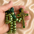 A hand holds a green and gold St. Benedict Rosary against a draped beige satin background.