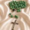 A green faceted bead rosary with a silver-tone crucifix and centerpiece, arranged on draped satin fabric.