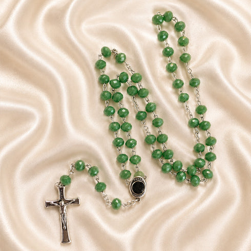 A green beaded rosary with a silver crucifix and centerpiece, arranged on a cream satin fabric background.