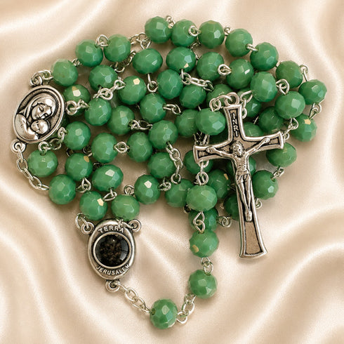 A green rosary with silver-tone metal links, a crucifix, a round medallion, and a centerpiece, on a cream satin fabric background.