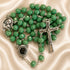 A green rosary with silver-tone metal links, a crucifix, a round medallion, and a centerpiece, on a cream satin fabric background.
