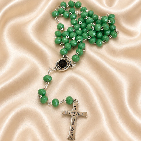 A green rosary with a silver-tone cross pendant engraved with 'JESUS', on a draped beige satin fabric background.