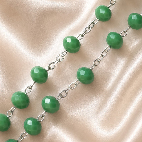 A close-up of a beaded chain featuring green beads separated by silver-toned metal links, laid across a draped satin fabric.