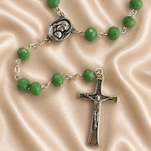 A green bead rosary with a silver crucifix and centerpiece medal, displayed on a beige satin fabric background.