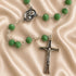A green bead rosary with a silver crucifix and centerpiece medal, displayed on a beige satin fabric background.