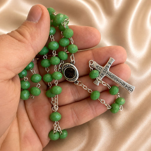 A close-up of a hand holding a green beaded rosary with silver-tone chain links, a round black centerpiece medallion, and a silver cross pendant.