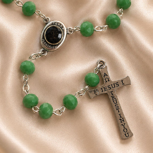 A close-up of a rosary with faceted green beads, a silver-tone centerpiece medallion, and a silver-tone cross engraved with 'JESUS', on a beige satin fabric background.