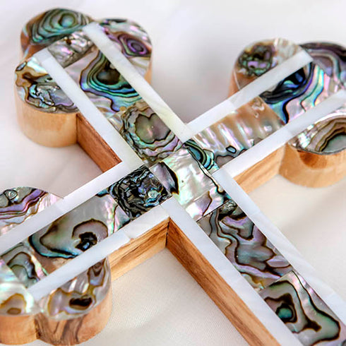 Promotional image of a detail of a handcrafted budded olive wood cross with green-white mother of pearl accents, placed on a white background with rose petals beside it.