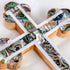 Promotional image of a detail of a handcrafted budded olive wood cross with green-white mother of pearl accents, placed on a white background with rose petals beside it.