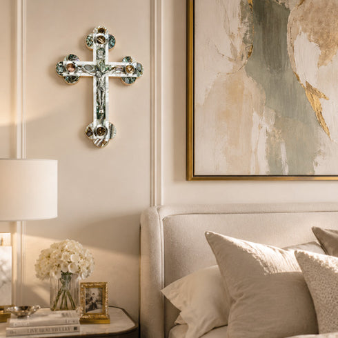 Decorative cross on a wall above a bed with a lamp and artwork.