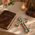 Holy Bible, decorative cross, and lit candle on a table with flowers and religious artwork.