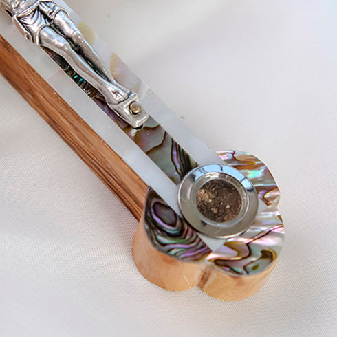 Zoomed detail image of the bottom of an olive wood budded cross with holy soil capsule adorned with a silver crucifix and colourful mother of pearl, laid on a clean white surface.