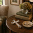 Decorative cross on a wooden table with books and a vase of flowers in the background.