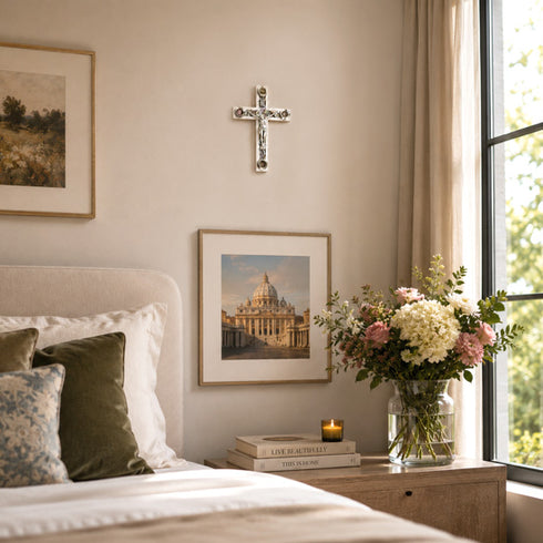 Neatly made bed with decorative pillows, framed picture, cross on wall, and flowers on nightstand.