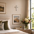 Neatly made bed with decorative pillows, framed picture, cross on wall, and flowers on nightstand.